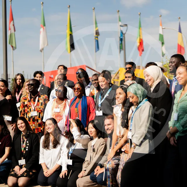 A large group of young World Food Forum participants posing in front of a row of flags under a blue sky.