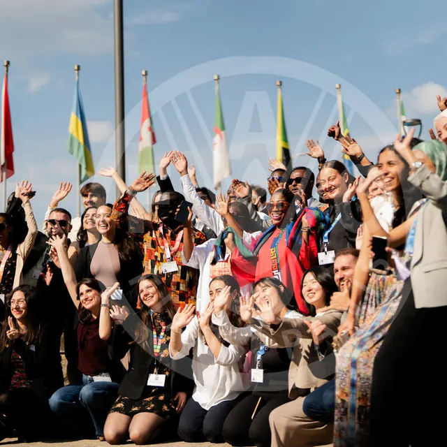Young World Food Forum participants waving and smiling in front of the flags, in bright sunshine.