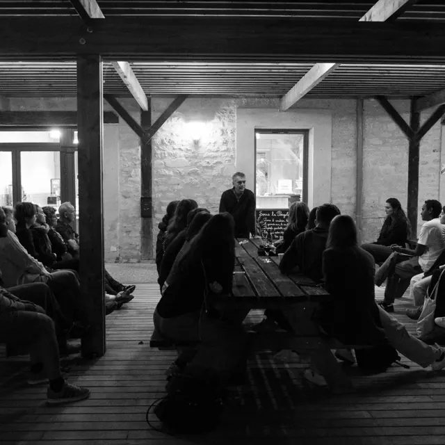 An evening talk in black and white: participants seated at long tables under a pergola listen to a standing speaker.
