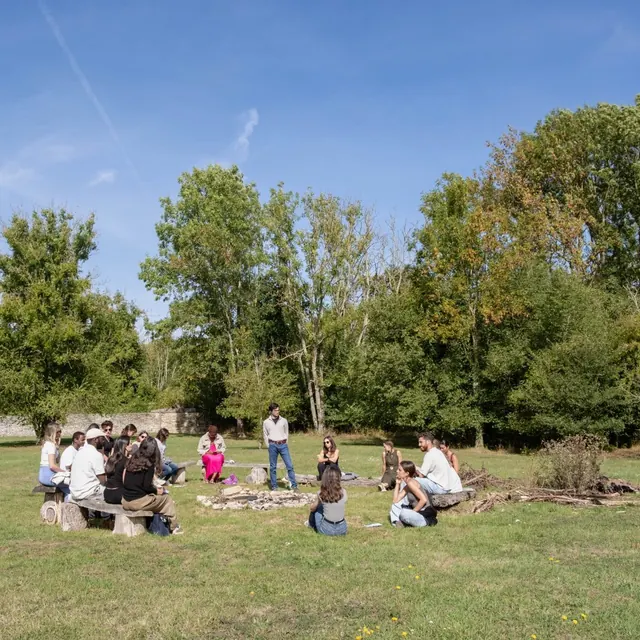 Participants seated in a circle on wooden benches in the middle of a lawn, around a stone fire pit, with a line of trees behind.